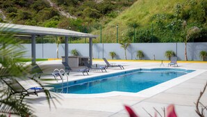 Una piscina con sillones de descanso y una zona cubierta, rodeada de vegetación y un paisaje montañoso.