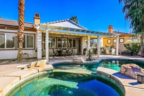A pool with a patio area, a covered seating area, and a house with a chimney.