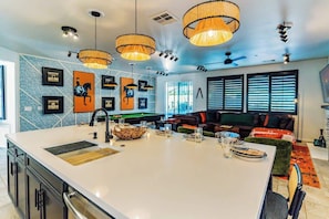 Uma cozinha moderna com ilha central, luminárias pendentes e mesa de bilhar ao fundo.