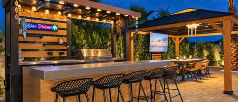 An outdoor bar area with a grill, bar stools, and a neon sign.