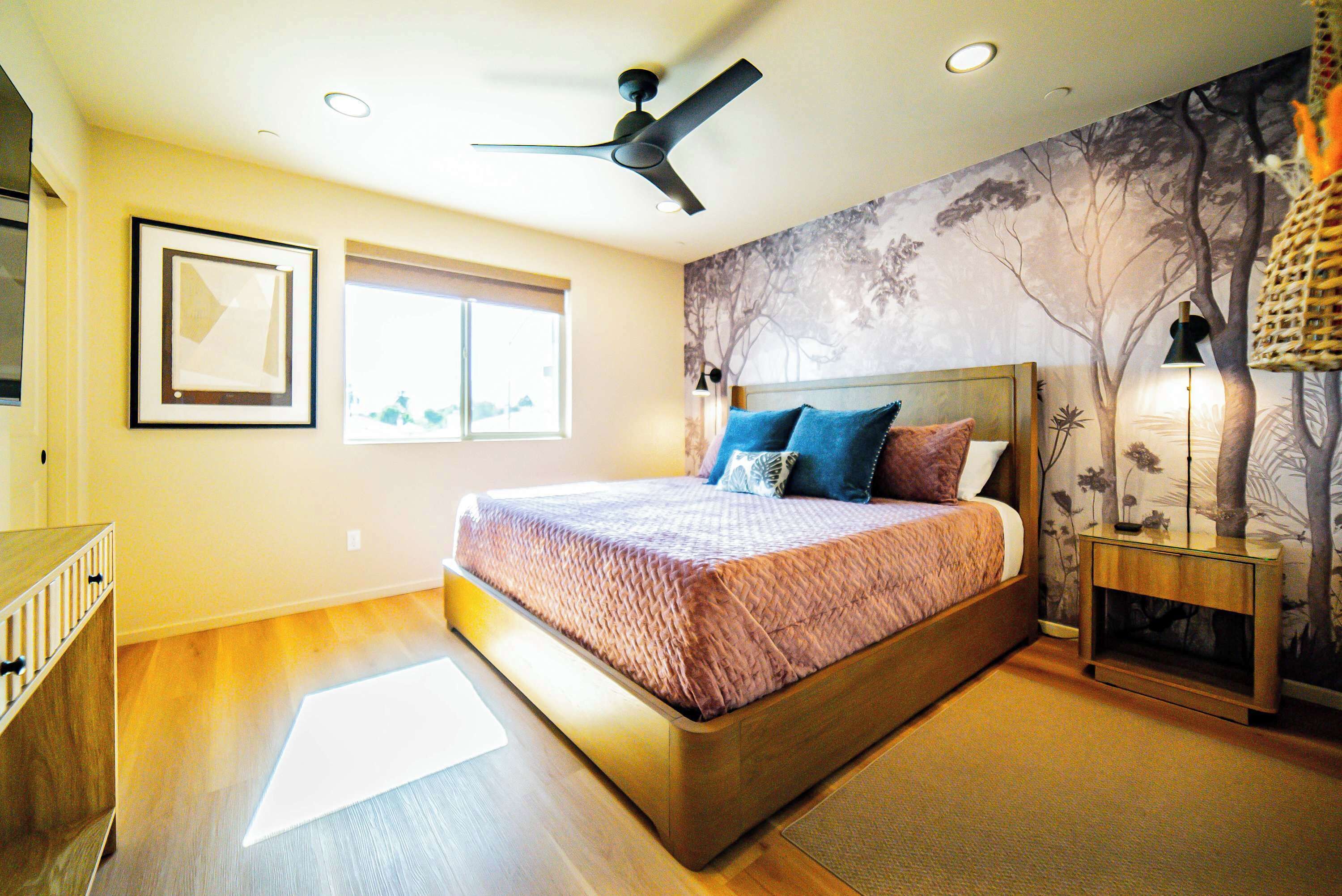 A bedroom with a bed, a ceiling fan, a window, and a nightstand with a lamp.