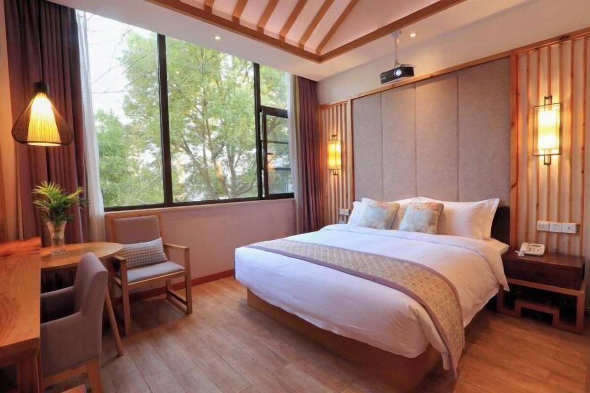 A hotel room with a bed, bedside table, lamp, chair, and a large window.