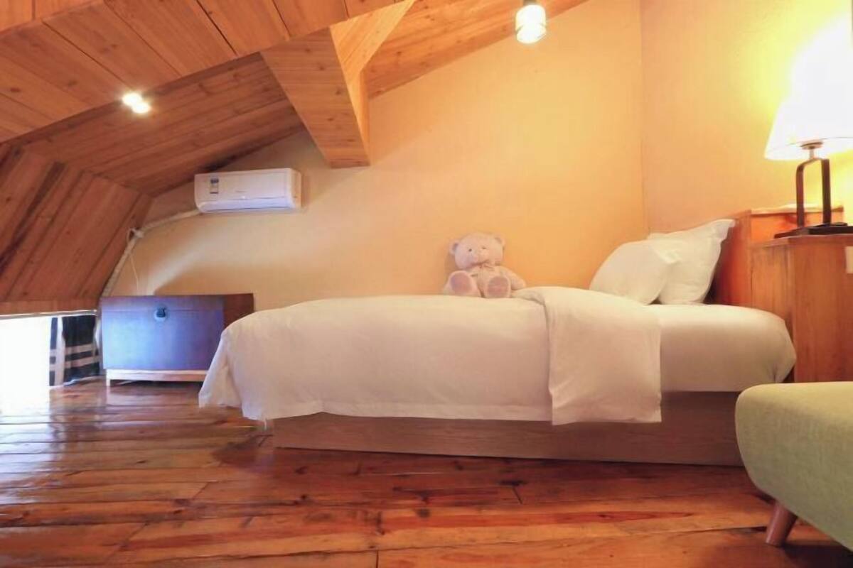 A cozy bedroom with a wooden ceiling, a bed with white bedding, a teddy bear on the bed, and a nightstand with a lamp.