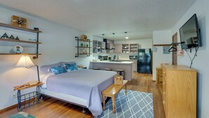 A compact bedroom with a bed, bedside table, and a small desk with a lamp. A kitchenette with a fridge, stove, and sink is visible in the background. - Mtn Views: Waterfront Red River Studio (Red River)