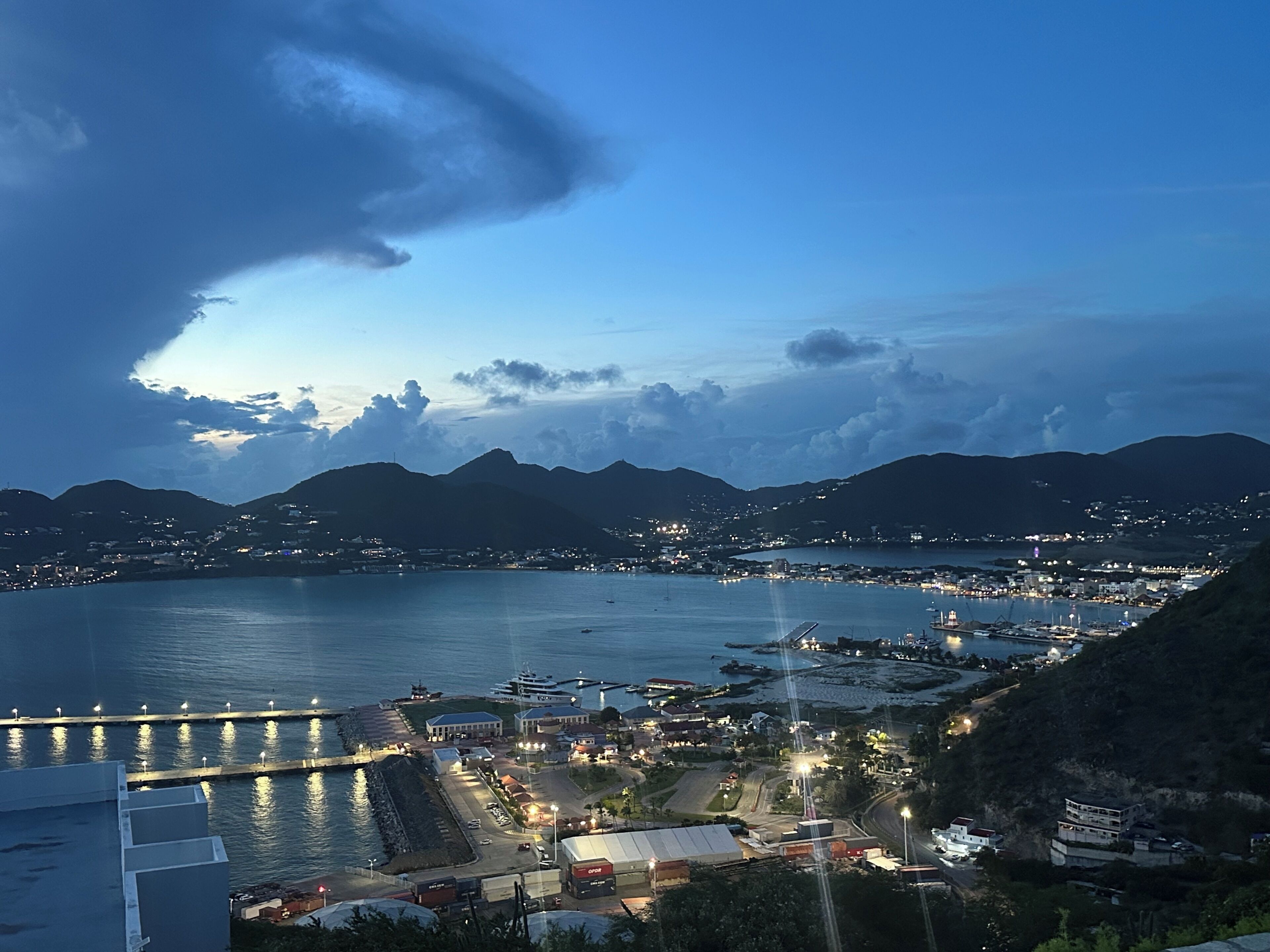 A coastal town at dusk with a marina, buildings, and mountains in the background.