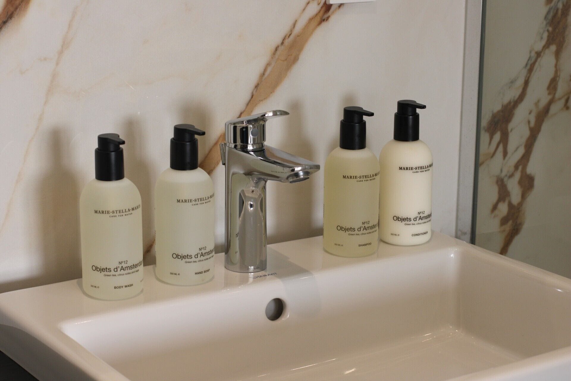 A bathroom sink with four bottles of Marie-Stella-Mare body wash and shampoo, a modern faucet, and a marble backsplash.