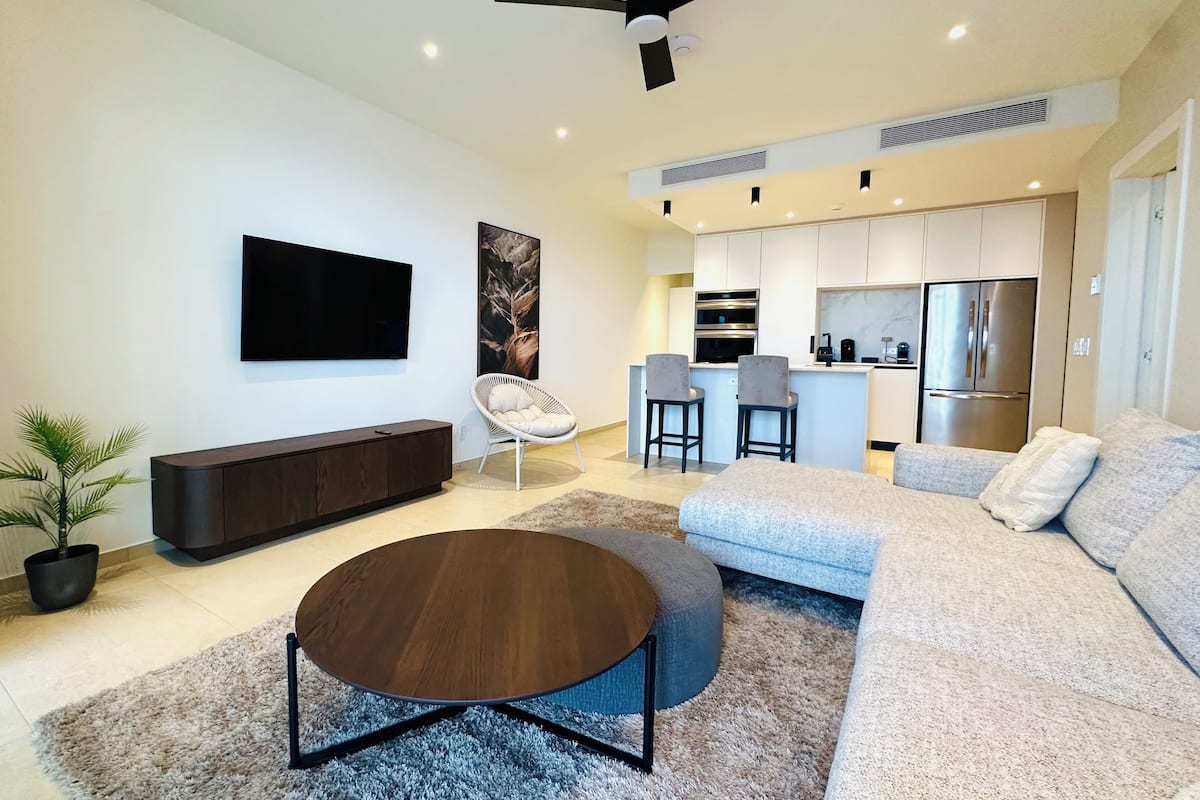 Una sala de estar moderna con un televisor de pantalla plana, una mesa redonda de centro, un sofá y un comedor con una cocina al fondo.
