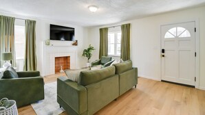 A living room with a green sofa, a flat-screen TV, a fireplace, and a door with a window. - Walk to Ritter Park: Renovated Home Near Marshall! (Huntington)