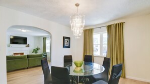 A modern living room with a glass dining table, black chairs, a green sofa, a chandelier, and a TV mounted on the wall. - Walk to Ritter Park: Renovated Home Near Marshall! (Huntington)