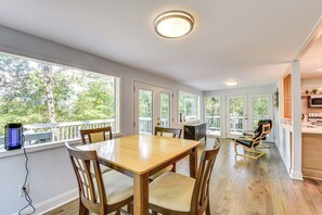 Un comedor amplio con una mesa y sillas de madera, ventanas grandes que ofrecen vistas al exterior, y una cocina visible al fondo.