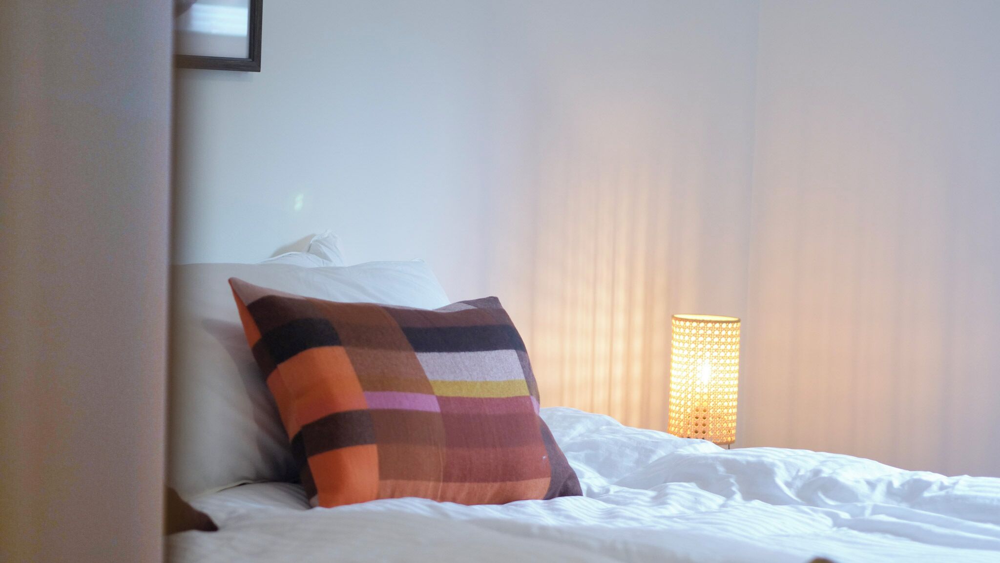 Ein ordentlich bezogenes Bett mit einem farbenfrohen Kissen in Streifen und einer Wandlampe.