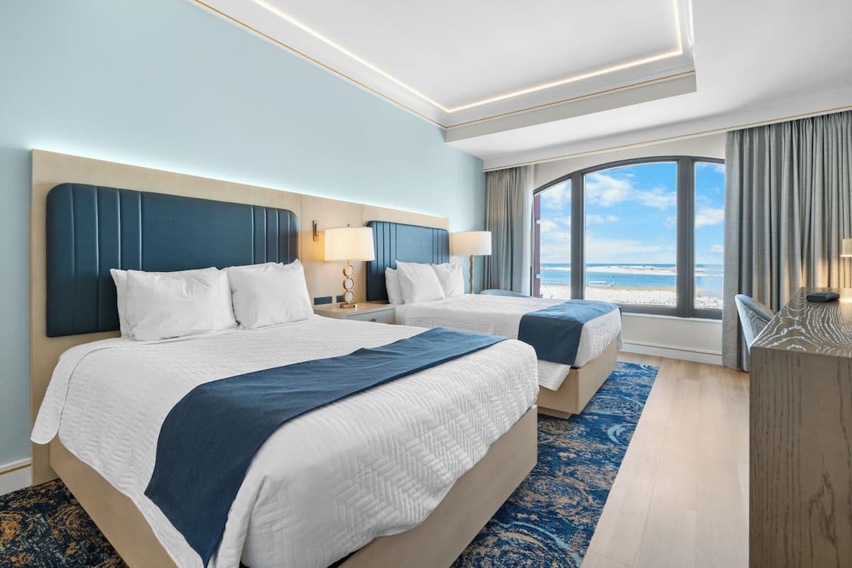 A hotel room with two beds, a large window with a beach view, and a desk with a lamp.