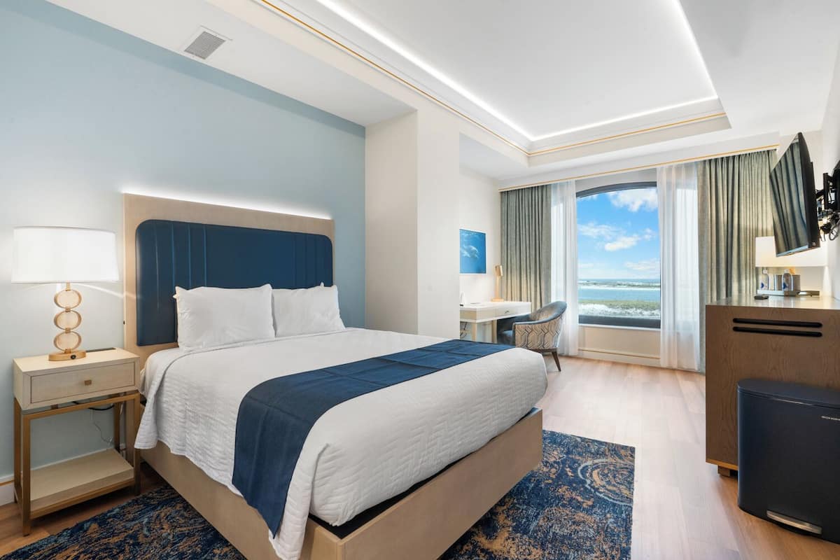 A hotel room with a large bed, a desk, a chair, a TV, and a view of the ocean.