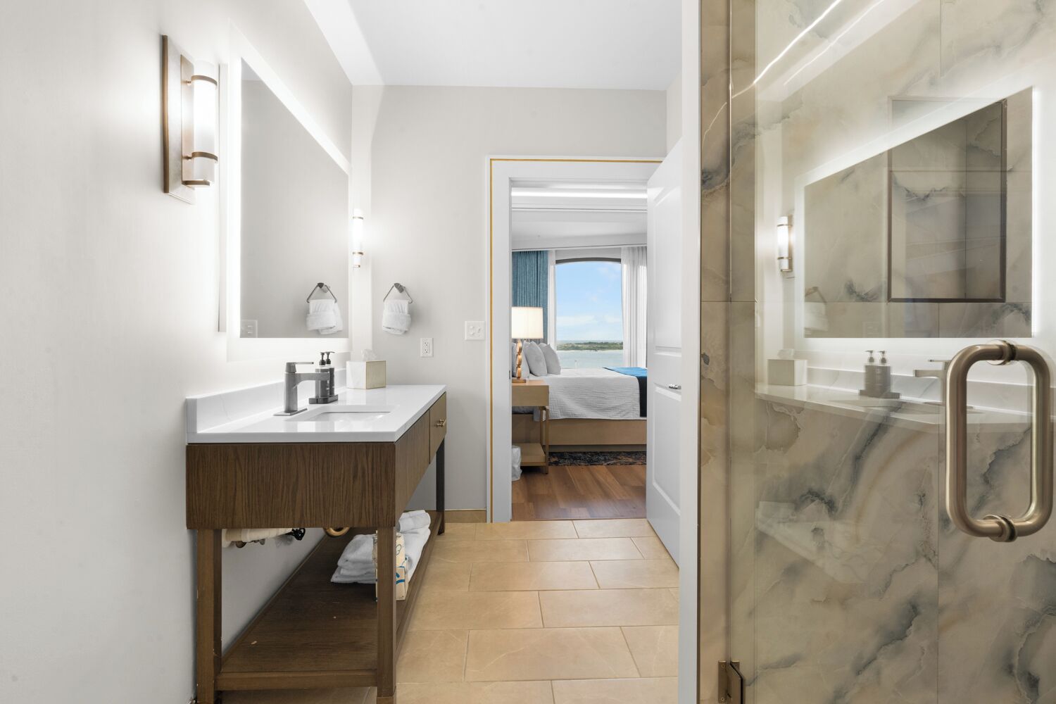 Une salle de bain moderne avec une douche en marbre, un meuble-lavabo en bois et une vue sur une chambre à coucher avec un lit et une fenêtre donnant sur la mer.