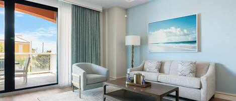 Una sala moderna con sofá, sillón y una mesa de centro, una ventana grande con cortinas y un cuadro de una playa.