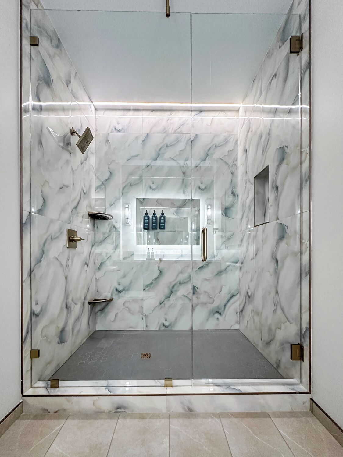 A glass-enclosed shower with marble walls, a built-in shelf with toiletries, and a metal handle.