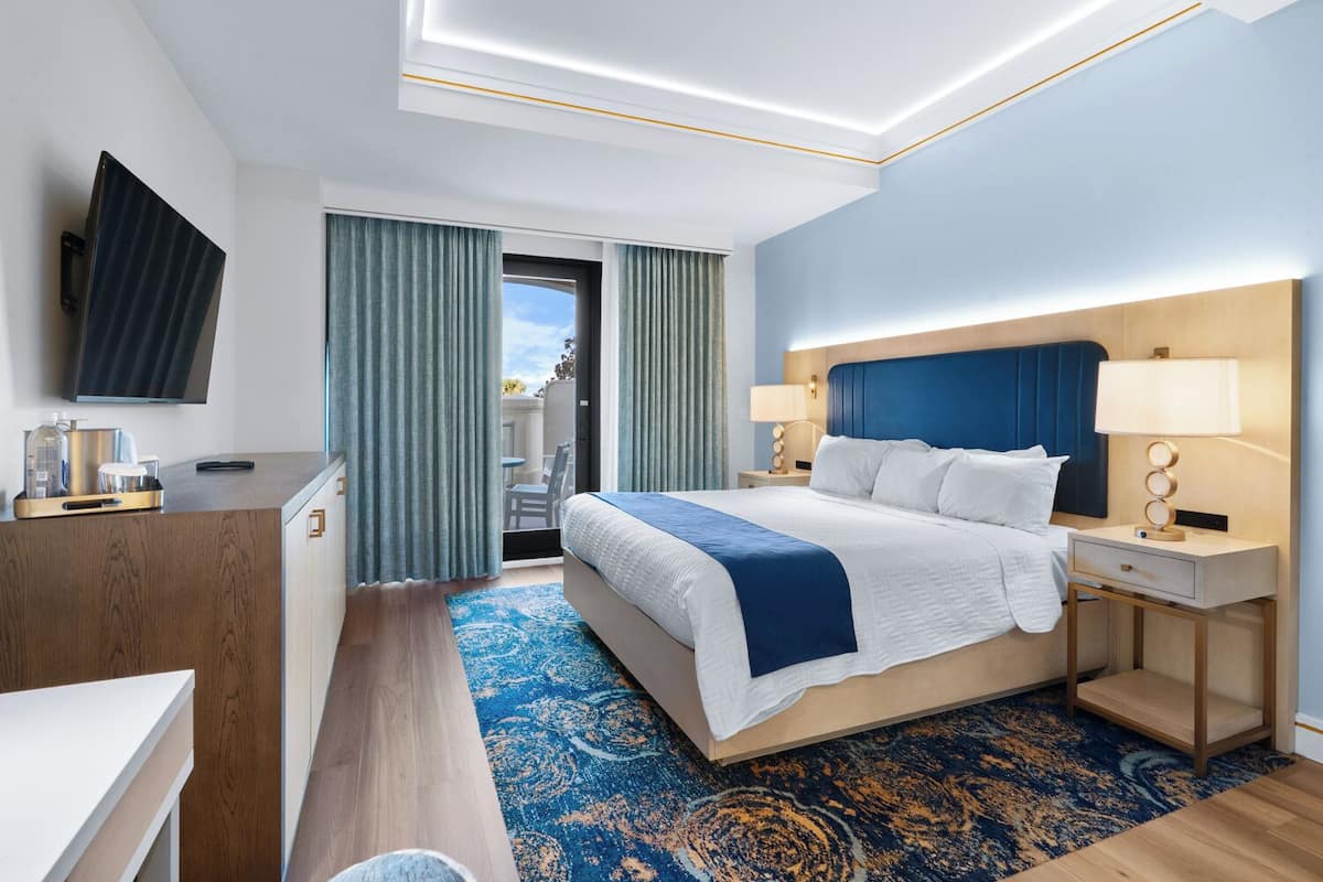 A modern hotel room with a large bed, a desk, a TV, and a balcony with curtains.