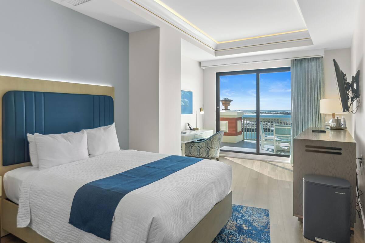 A hotel room with a large bed, a desk, a chair, a small trash bin, and a view of the city.