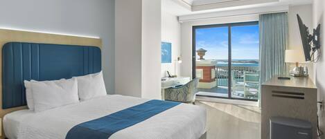 A hotel room with a large bed, a desk, a chair, a small trash bin, and a view of the city.