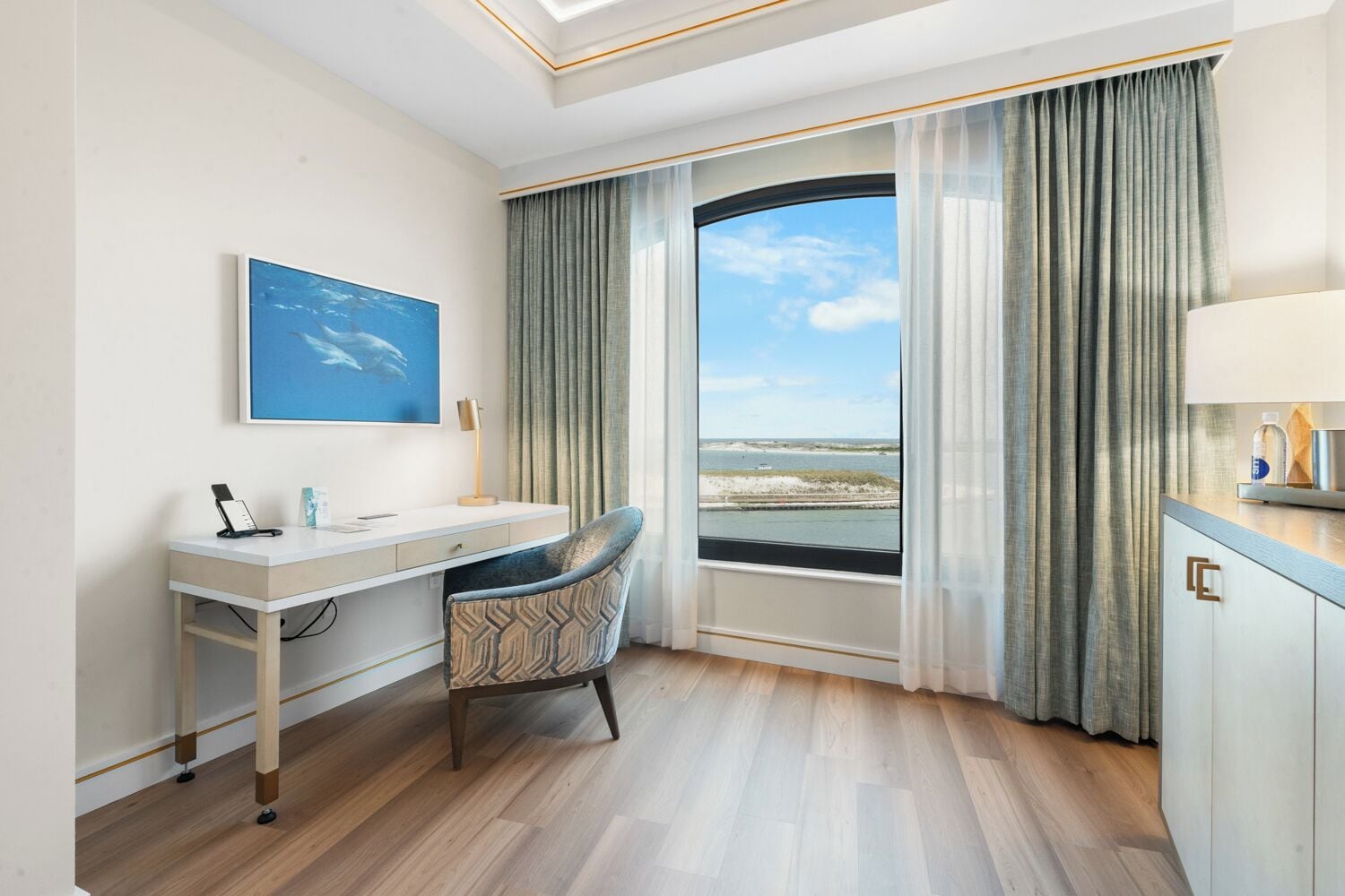 A hotel room with a desk, chair, and a large window with a view of the beach.