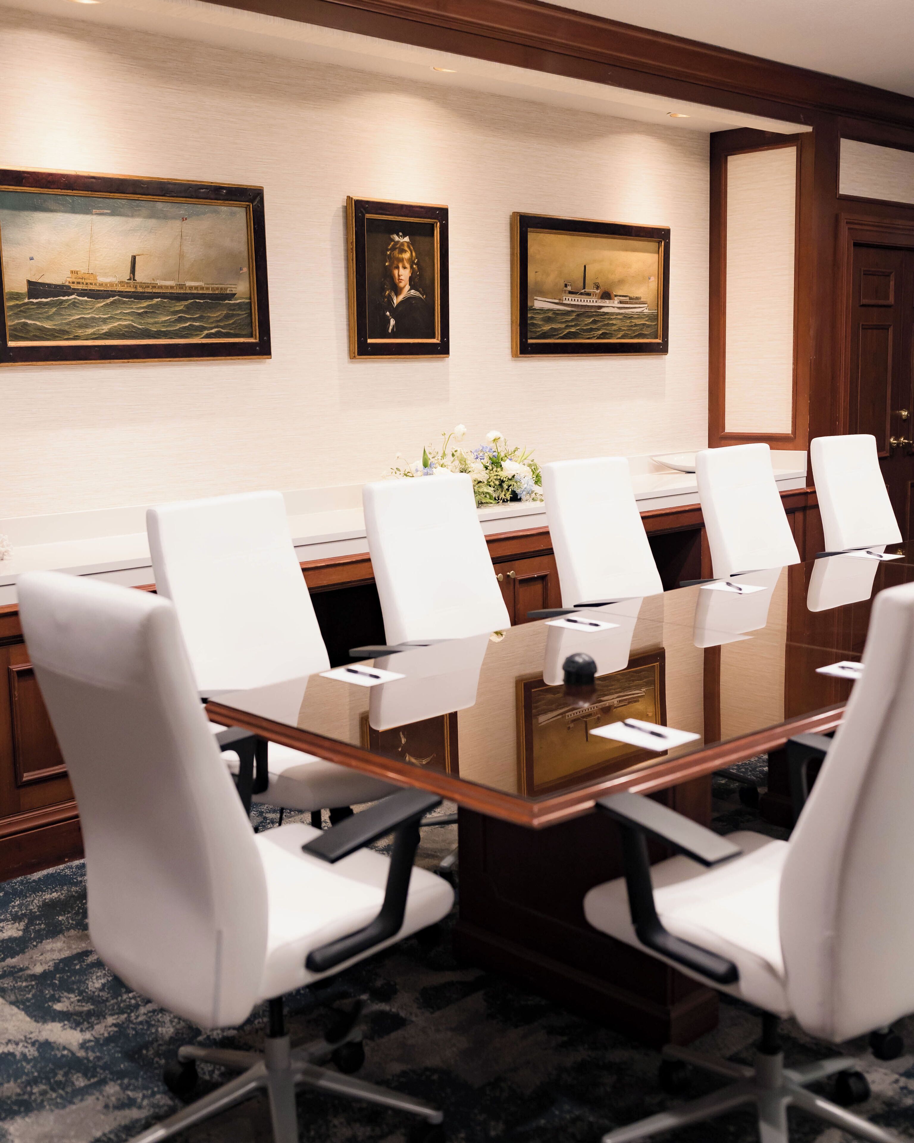 A conference room with a glass table, white chairs, and framed maritime paintings on the wall.