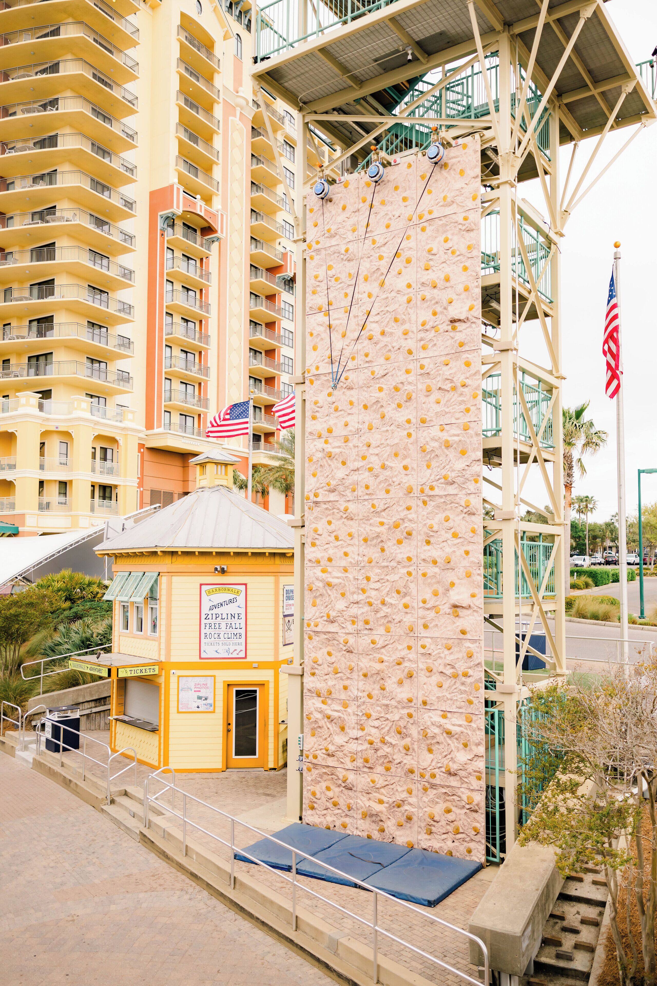 A rock climbing wall with an American flag, a ticket booth, and a building with balconies in the background.