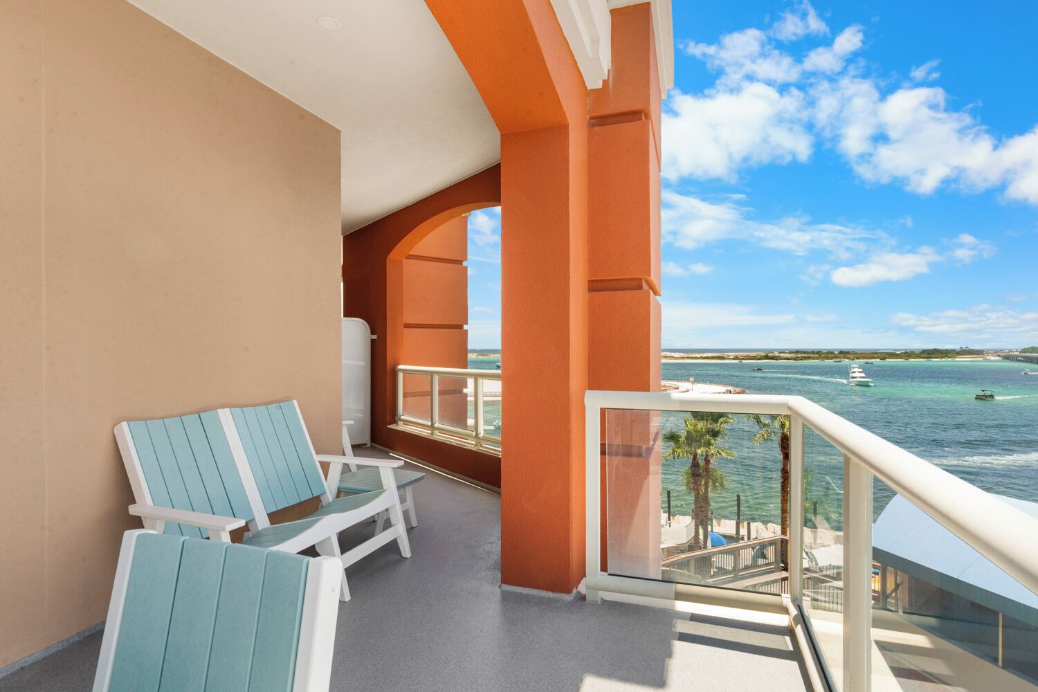 A balcony with two light blue chairs and a white table, overlooking a clear blue sea with boats and a distant shoreline.
