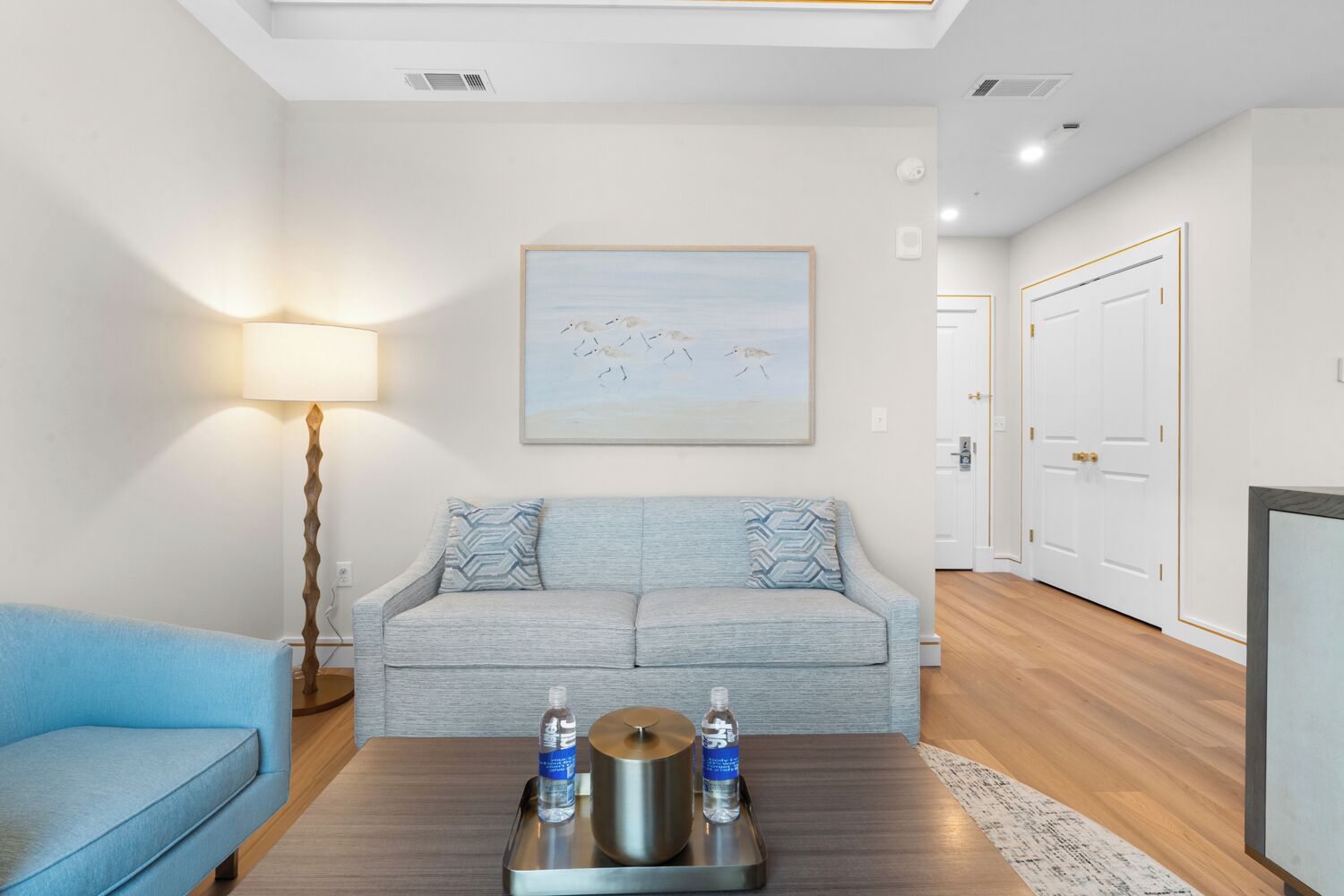 A modern living room with a light blue sofa, a matching armchair, a coffee table with water bottles, a floor lamp, and a painting of birds on the wall.