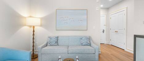 A modern living room with a light blue sofa, a matching armchair, a coffee table with water bottles, a floor lamp, and a painting of birds on the wall.