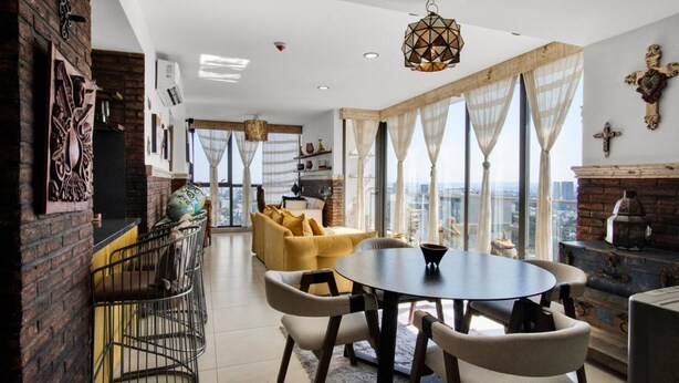 Una sala moderna con un sofá amarillo, una mesa de comedor redonda y vistas a la ciudad a través de grandes ventanales.