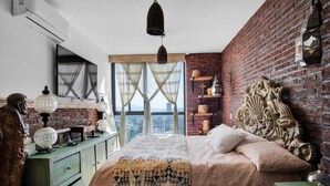 Un dormitorio con pared de ladrillo, cama con ropa de cama estampada, cómoda y un ventanal con cortinas.
