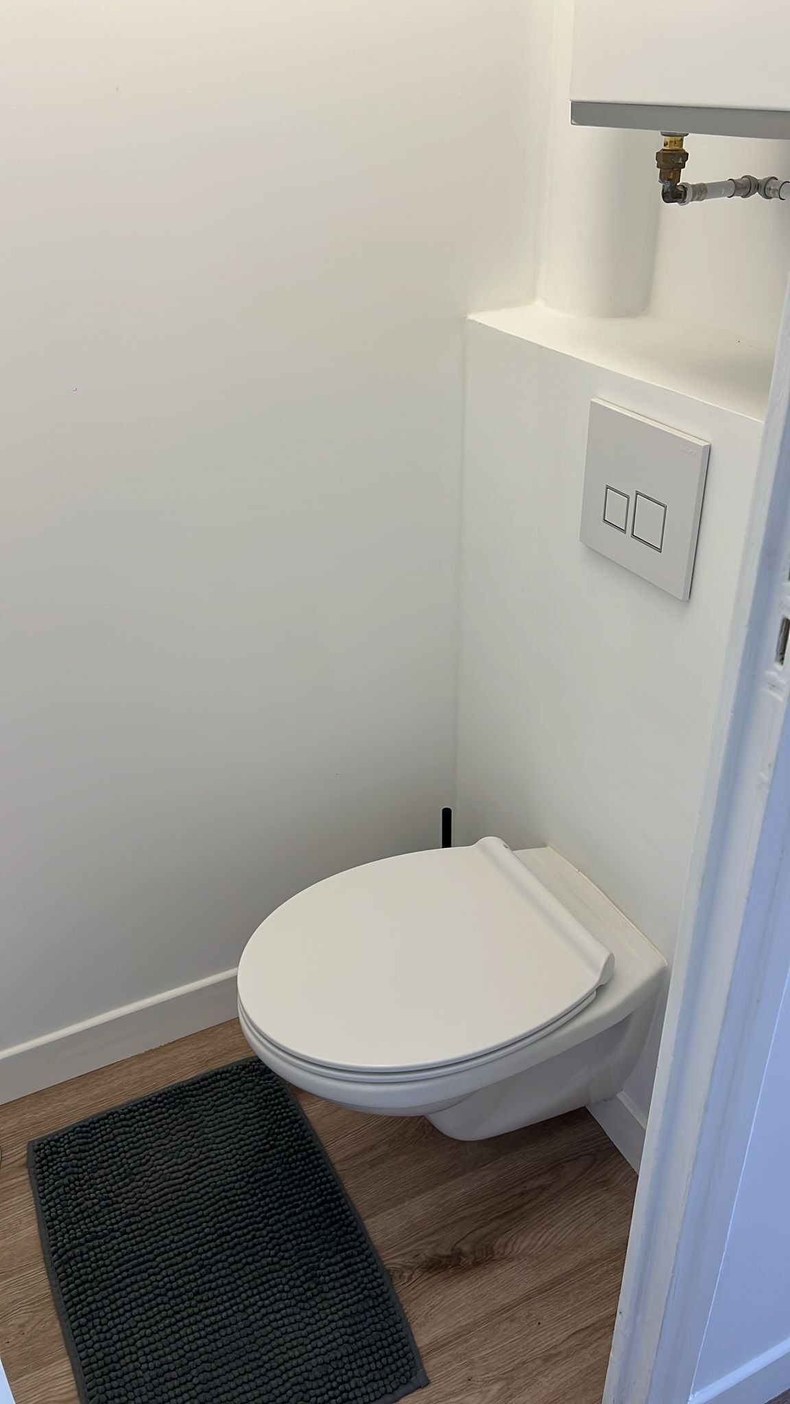 Et lille, hvidt toilet med lukket låg, en sort, struktureret måtte foran og et skyllebræt på væggen.