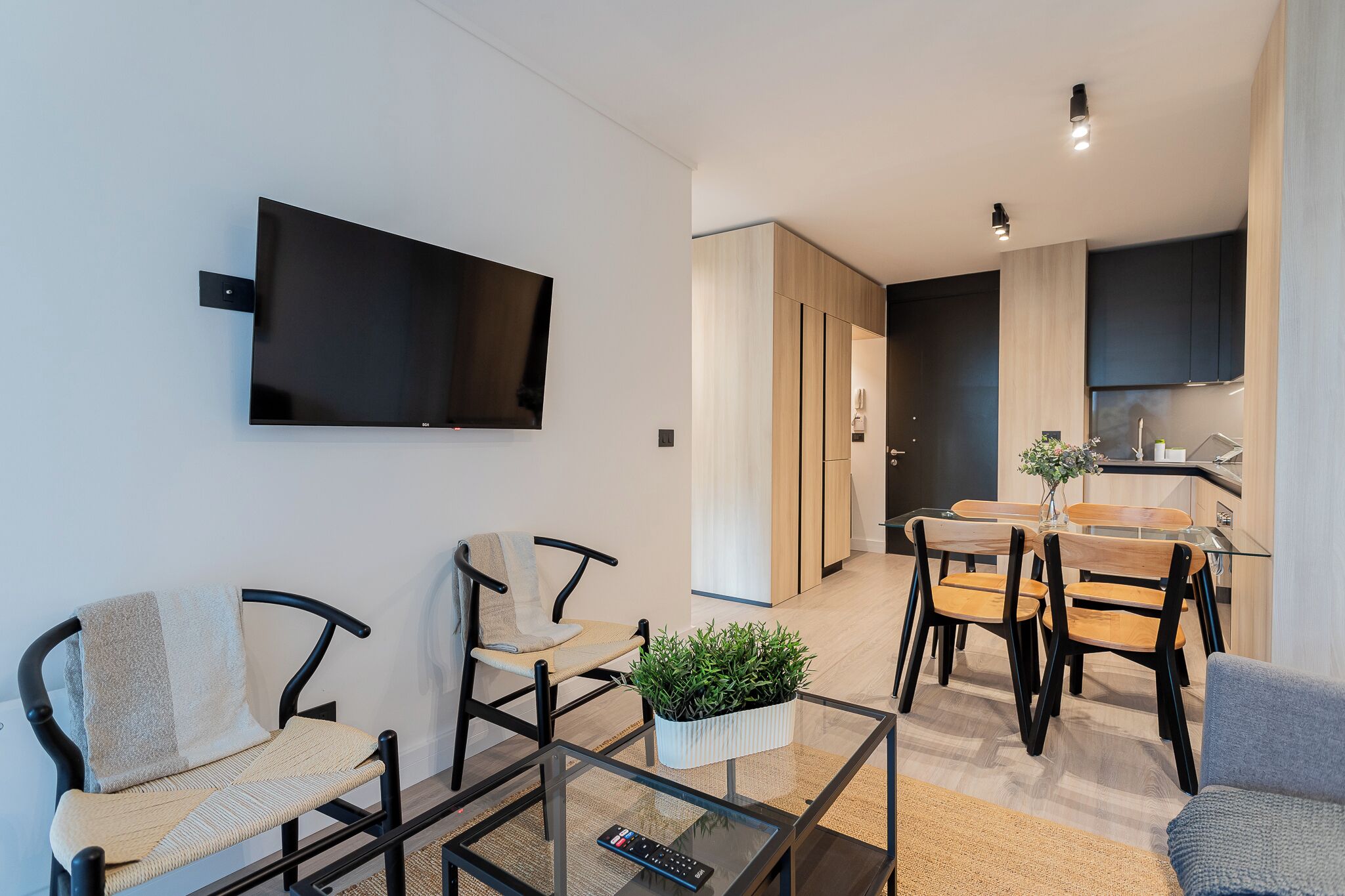 A modern living room with a flat-screen TV, two chairs, a glass coffee table with a remote, and a dining area with wooden chairs and a small plant.