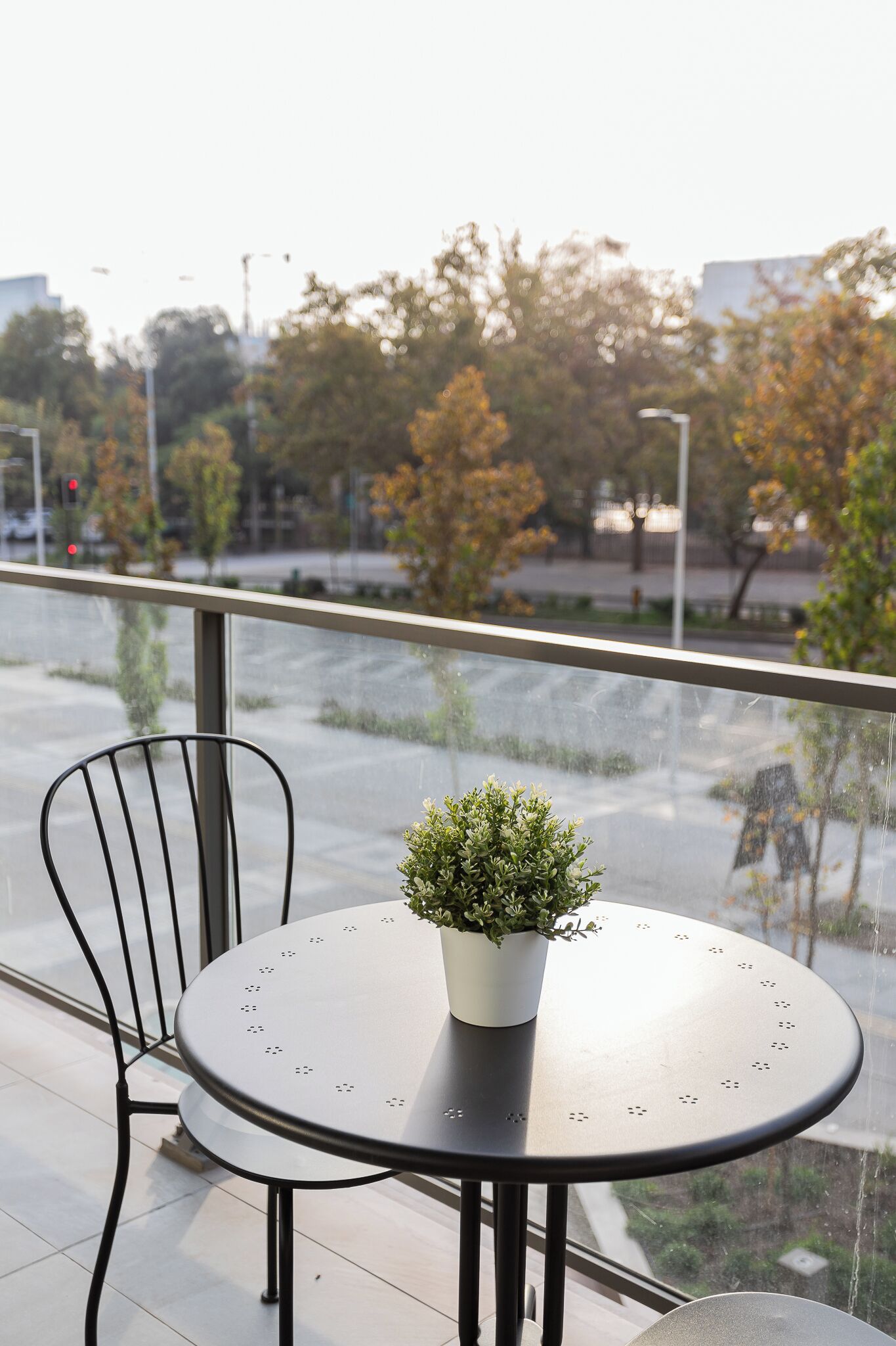 A round metal table with a potted plant on a balcony with a view of trees and a street.