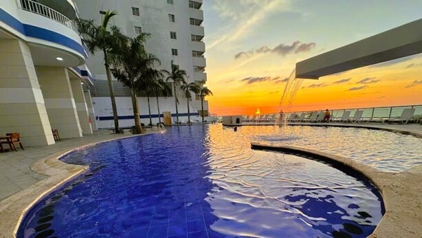 A swimming pool with a sunset view, surrounded by a building with balconies and palm trees.