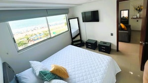 A bedroom with a bed, a TV, a window with a view of the beach, and a door leading to another room.