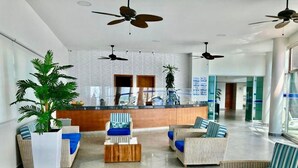 A spacious lobby with a reception desk, multiple seating areas with blue cushions, ceiling fans, and large potted plants.