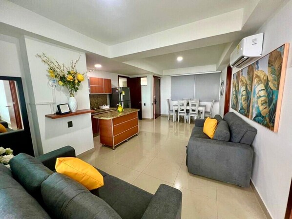 A modern living room with a grey sofa and ottoman, a dining area with a white table and chairs, and a kitchen with wooden cabinets and a refrigerator.