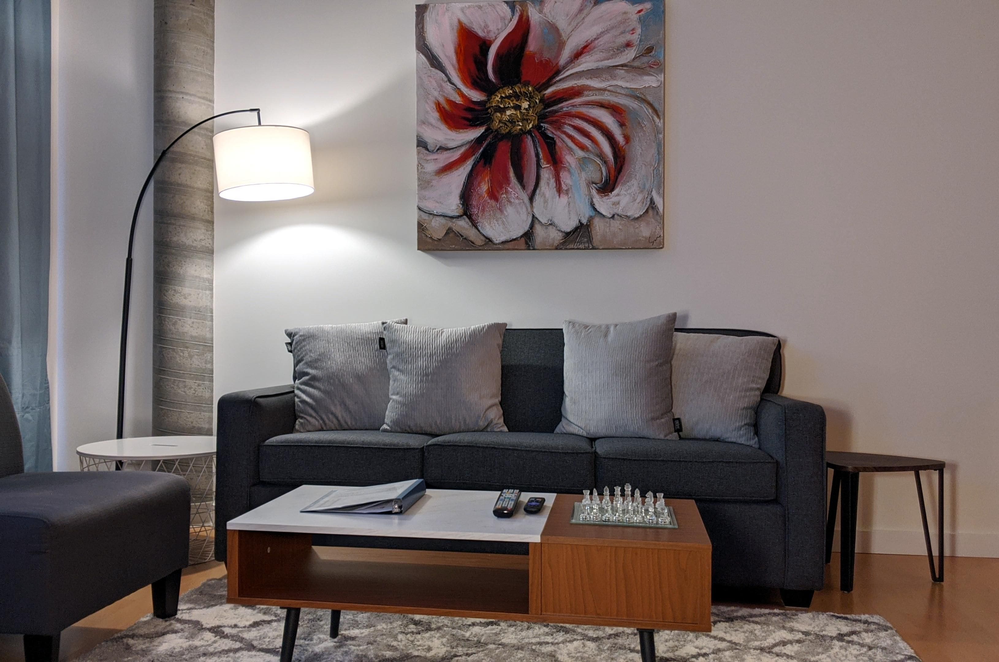 A modern living room with a grey sofa, a coffee table, a chess set, and a painting of a flower on the wall.