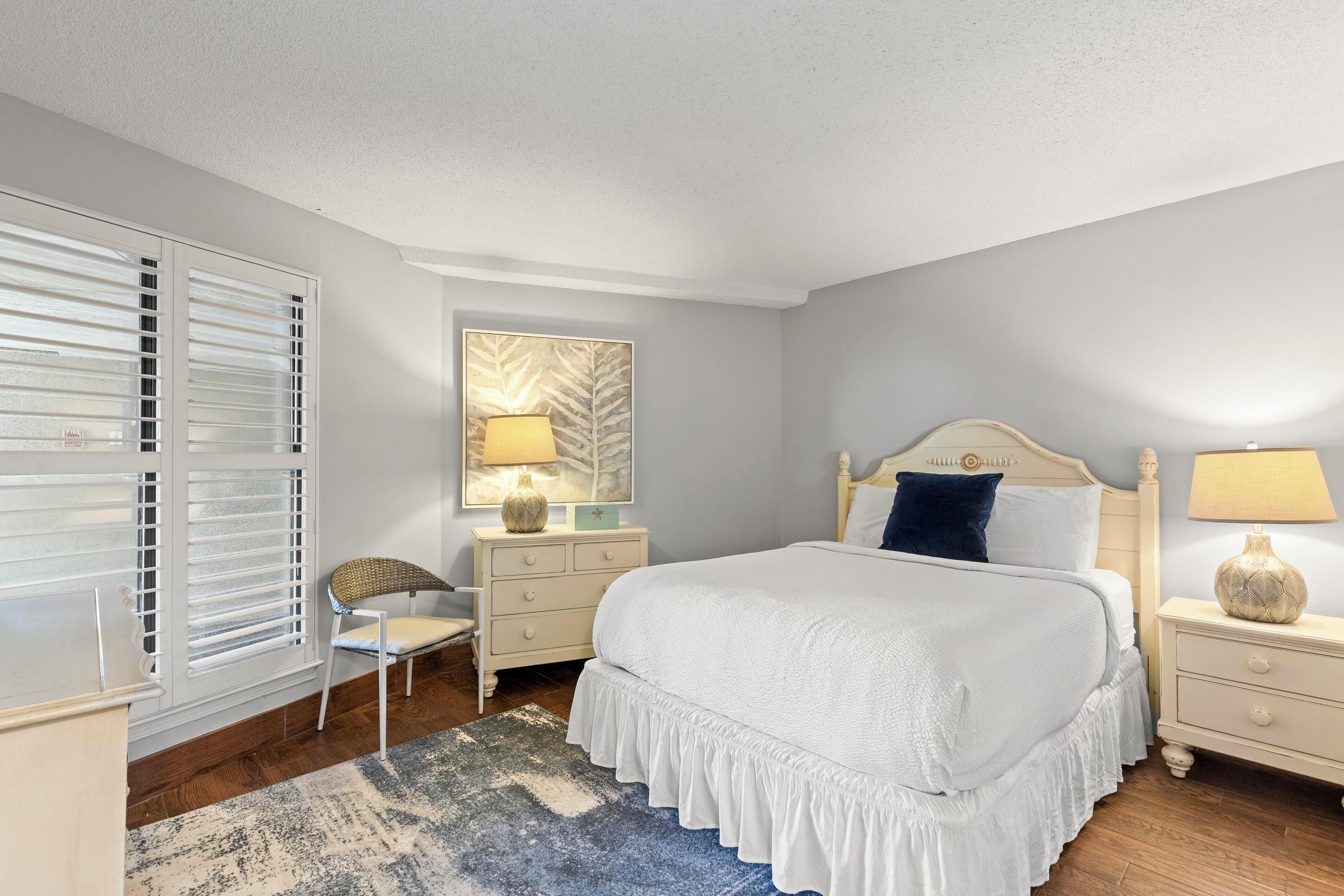 A bedroom with a large bed, a nightstand, a chair, a lamp, and a window with blinds.