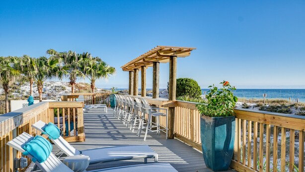 Een houten terras met lounge fauteuils en een pergola met uitzicht op een strand.