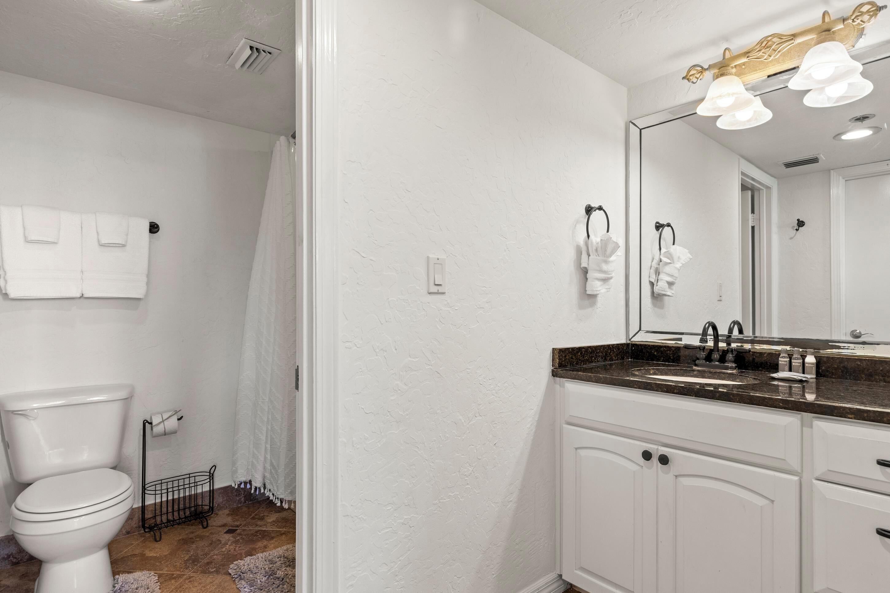 A bathroom with a white toilet, a sink with a dark countertop, and a mirror with lights above it.