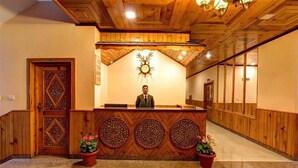 A reception desk with a clock above it, wooden paneling on the walls, and potted plants on either side. - Luxe Signature  (Manali)