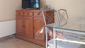 Een eetgedeelte met een glazen tafel, metalen stoelen, een houten kast met een televisie erboven, en twee ingelijste foto's aan de muur.
