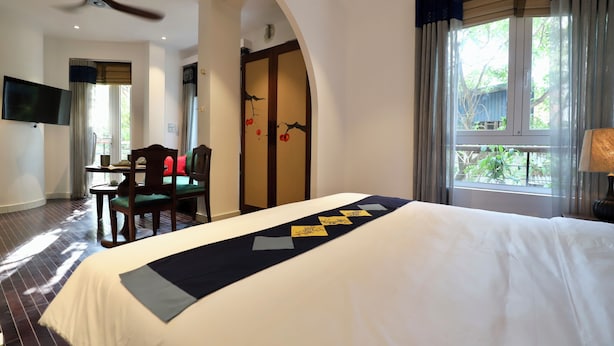 A hotel room with a large bed, a ceiling fan, a TV mounted on the wall, a dining table with chairs, and a view of the outdoors through the window. - Tranquil Truc Bach (Hanoi)