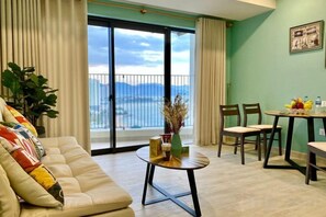 A living room with a sofa, a coffee table, and a dining area with chairs. There is a large window with a view of the sea and mountains. - MASTER Gold Coast Ocean View (Nha Trang)