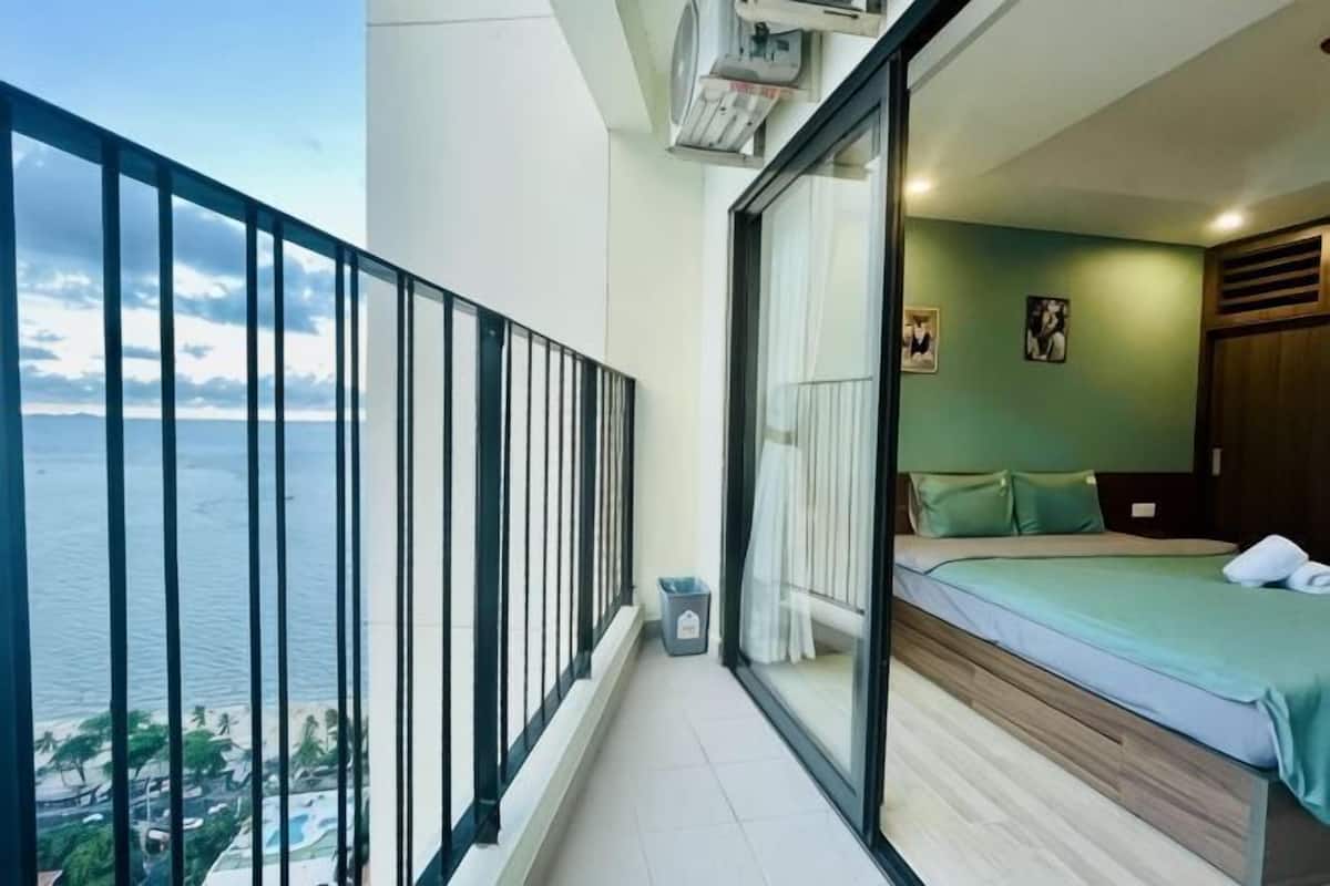 A balcony with a view of the sea, a bed with a green bedspread, and a small trash bin.