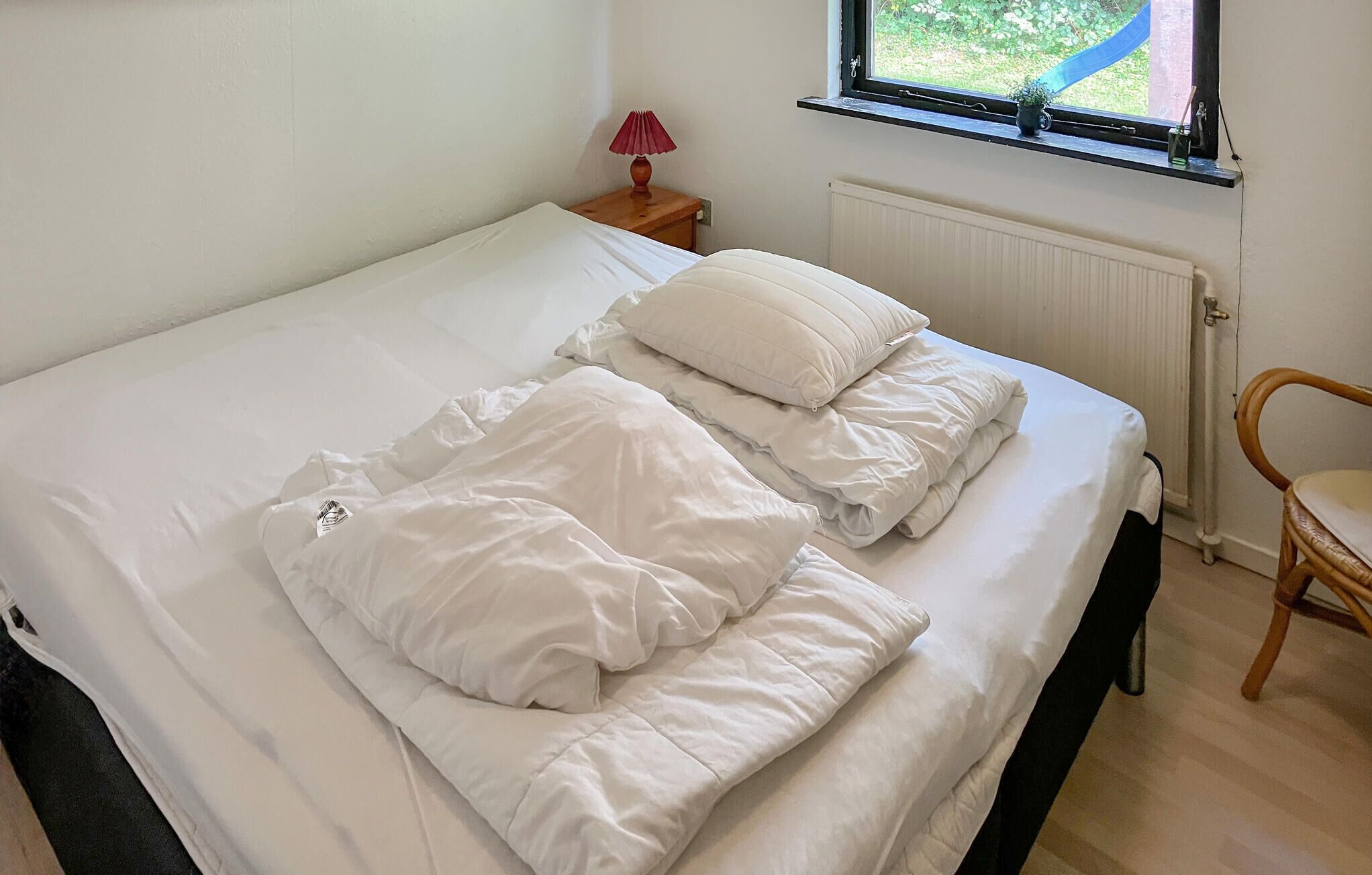 Un lit bien fait, avec du linge de lit blanc, dans une pièce dotée d’une fenêtre, d’une petite table avec une lampe et d’une chaise.