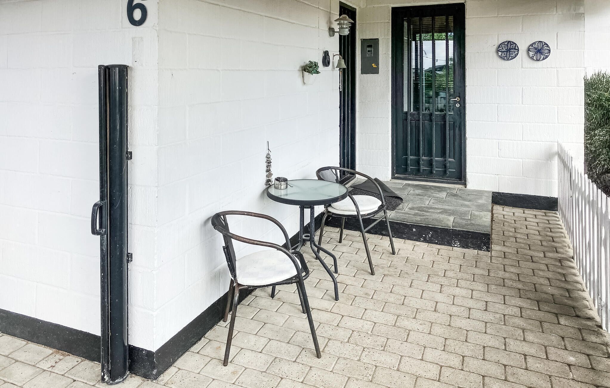 Un pequeño espacio al aire libre con dos sillas de metal y una mesa de vidrio, una puerta negra y una pared blanca con un letrero con el número '6'.