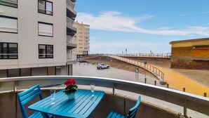 Un balcon avec des chaises bleues et une table, donnant sur une route où circulent des voitures et sur une plage.
